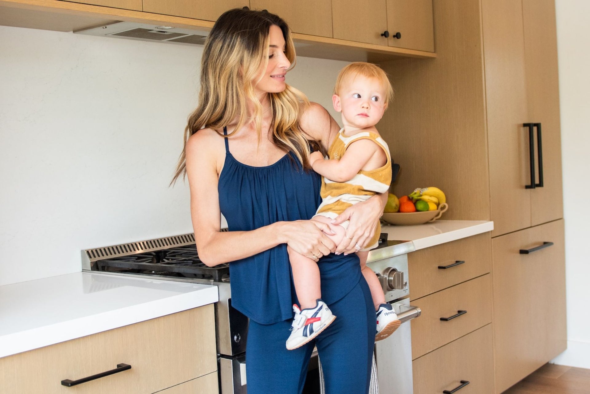 woman in navy JJ top and pants holding a baby in the kitchen.